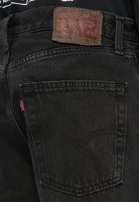 Black denim jeans featuring a brown leather patch with embossed logo. Standard five-pocket design with red tab on the back pocket.