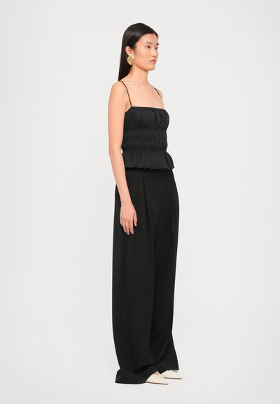 Black peplum top with gathered texture and adjustable spaghetti straps, paired with loose black trousers and cream-colored pointed shoes.
