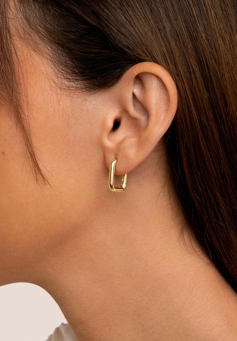 Pendientes de aro dorados con forma rectangular, textura suave y acabado pulido, usados en el lóbulo de la oreja de una modelo.