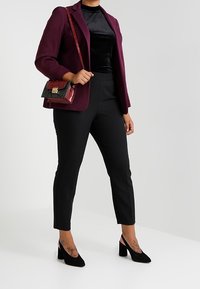 Bordeaux blazer met ribbelstructuur, zwart fluwelen top, slanke zwarte broek en zwarte hakken. Kleine handtas met rode en groene accenten.