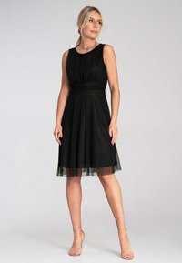 Schwarzes ärmelloses Kleid mit eng anliegendem Oberteil, gerafftem Detail und fließendem Tüllrock. Glatter Stoff mit transparenter Überlage am Saum.