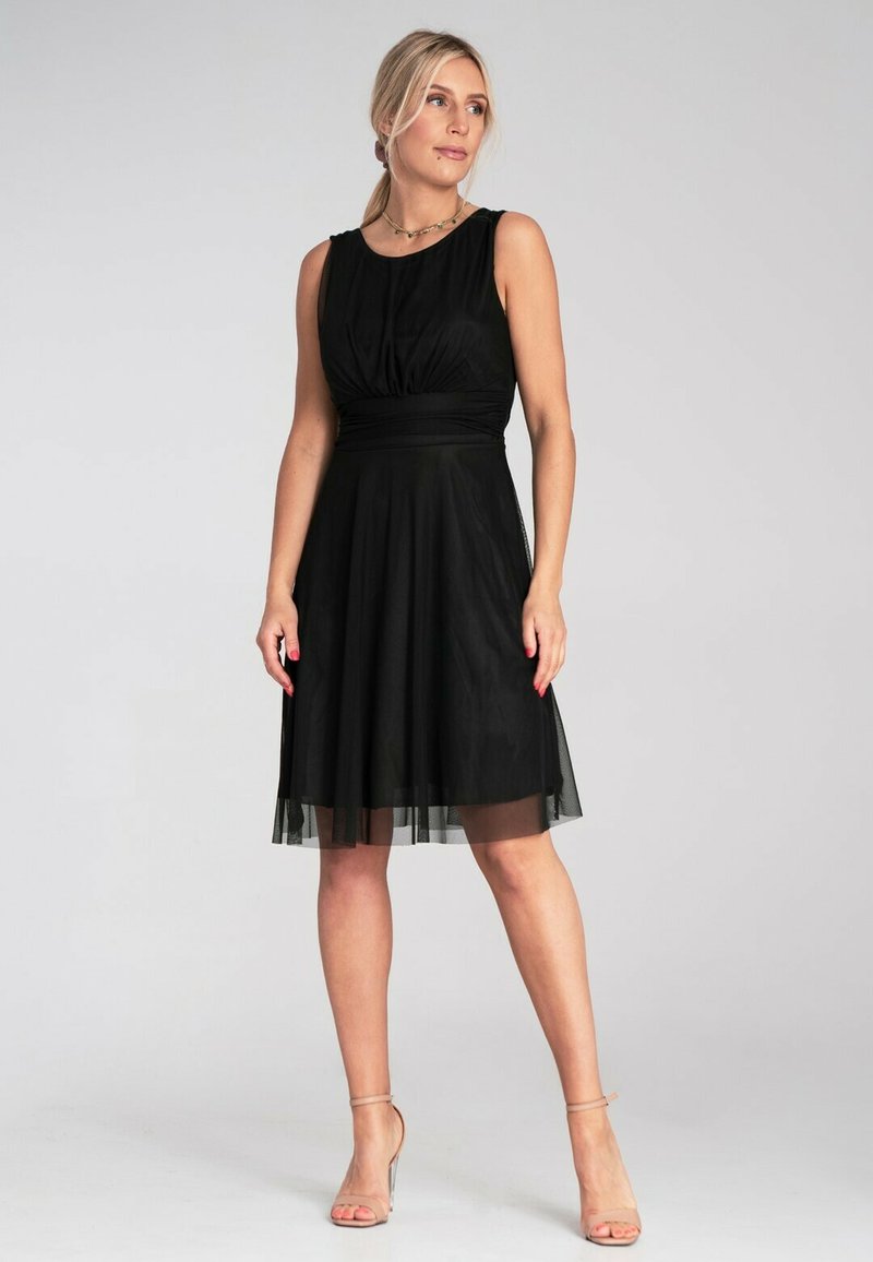 Schwarzes ärmelloses Kleid mit eng anliegendem Oberteil, gerafftem Detail und fließendem Tüllrock. Glatter Stoff mit transparenter Überlage am Saum.