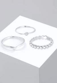 Drei silberne Ringe, die auf einer weißen Plattform ausgelegt sind: ein dünner Ring mit einem zentralen Stein, ein geformter Ring und ein Ring mit Perlen. Alle sind glatt und poliert.