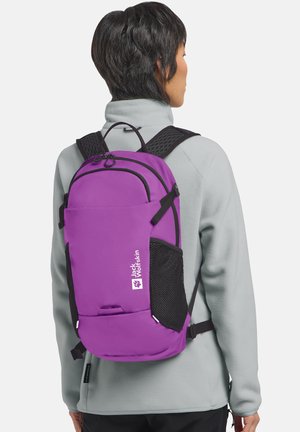 Persoon met een paarse Jack Wolfskin-rugzak met zwarte banden en zijvakken van gaas, gedragen over een lichtgrijs fleecejack, gezien van achteren.