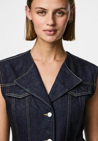 Mörk denim ärmlös klänning med krage, utrustad med vita knappar, kontrasterande gula stygn och dubbla bröstfickor.