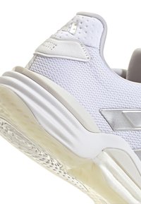 Chaussure de sport blanche avec tige en mesh, accents argentés, col rembourré et semelle en caoutchouc. Présente une finition texturée et des détails réfléchissants.