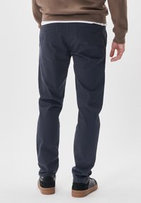 Pantaloni din bumbac bleu marin cu un croi slim, având două buzunare la spate și adidași cu talpă din cauciuc bej, văzuți din spate.