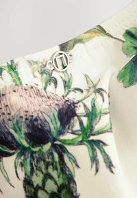 Stoff mit Blumenmuster in Grün und Lila, das Artischocken-Designs zeigt, mit einem silbernen, runden Verschlussdetail am Ausschnitt.