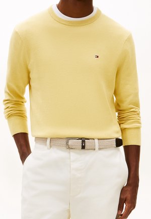 Mann trägt hellgelben Pullover mit kleinem Logo, weiße Hose und beigefarbenen geflochtenen Gürtel, Hände in den Taschen.