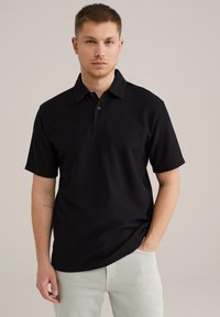 Črna polo majica s kratkimi rokavi iz rebrastega materiala, z klasičnim ovratnikom, zapenjanjem na tri gumbe in sproščenim krojem.