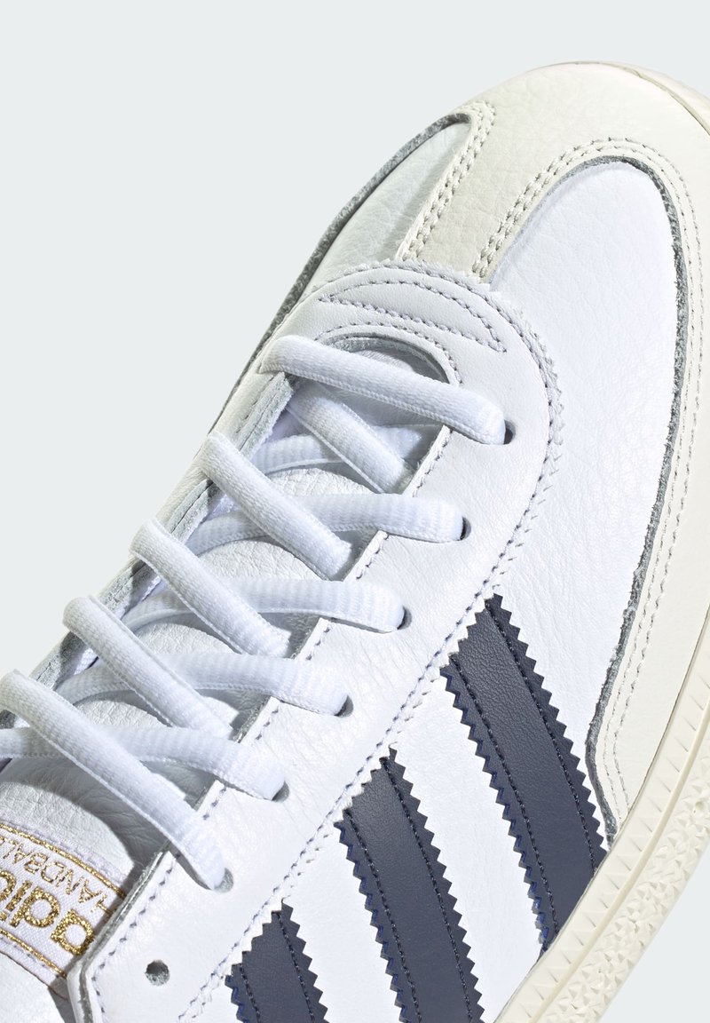 Białe skórzane sneakersy z trzema niebieskimi zygzakowymi paskami, okrągłymi białymi sznurowadłami i teksturowaną podeszwą z minimalnymi akcentami.