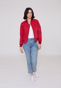 Rote Bomberjacke mit gerippten Manschetten und einer Reißverschlusstasche am Arm, kombiniert mit hellblauen Jeans und weißen Sneakers.