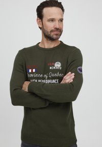 Tume roheline kootud sviiter ribidega mansettide ja ääristega. Omab erinevates värvides broderitud teksti ja tähist vasakul varrukal.