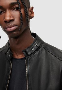 Schwarze Leder-Bomberjacke mit hohem Kragen, mit Druckknopfverschlüssen und silbernem Reißverschluss. Weiche Textur mit leicht glänzender Oberfläche.