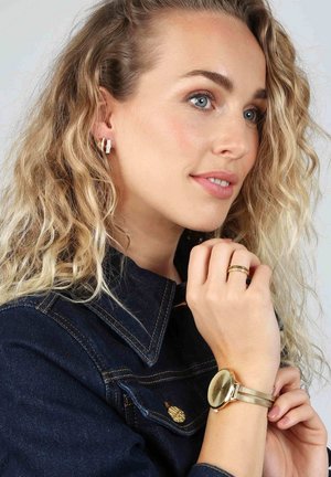Goldmesh-Armbanduhr mit rundem Zifferblatt, goldenen Akzenten und passendem dünnen Ring. Silberne Ohrhänger mit kleinen klaren Edelsteinen. Jeansjacke.