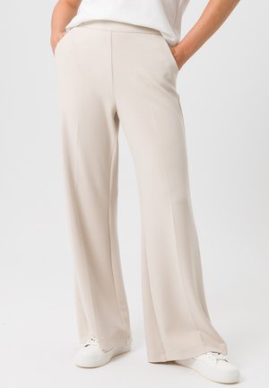 Beige weit geschnittene Hosen aus weichem Stoff, mit vorderen Taschen und einer glatten Textur. Kombiniert mit weißen Sneakers vor einem schlichten Hintergrund.