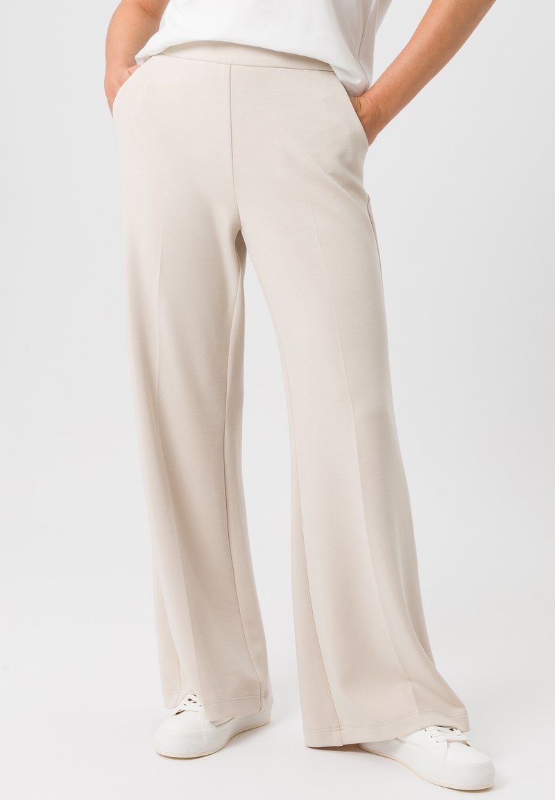 Beige wijde broek gemaakt van zachte stof, met voorzakken en een gladde textuur. Gecombineerd met witte sneakers op een effen achtergrond.