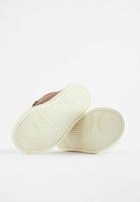 Chaussure marron avec semelle en caoutchouc blanc à motifs, arborant des designs géométriques. La tige est en cuir lisse, mettant en avant un style minimaliste.