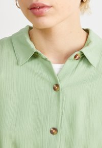 Chemise vert clair à col en tissu texturé avec deux boutons marron. Vues en gros plan mettant en avant le col et le tissu de la chemise.