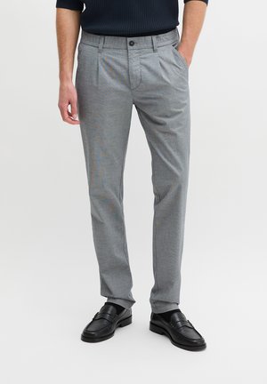 Mann trägt graue maßgeschneiderte Hose, schwarze Loafers und einen marineblauen Pullover, eine Hand in der Tasche, stehend vor weißem Hintergrund.