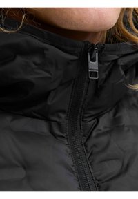 Schwarze Pufferjacke mit hohem Kragen, ausgestattet mit einem zentralen Reißverschluss und strukturiertem Stoff. Nahaufnahme des Reißverschlussbereichs und des Kragens.
