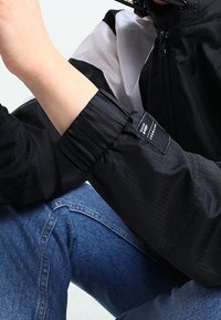 Person som bär en svart zip-up jacka med elastiska muddar och blå jeans, arm böjd med handen nära ansiktet mot en enkel bakgrund.