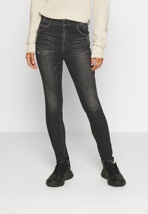 Osoba nosząca wyblakłe czarne obcisłe dżinsy, czarne masywne sneakersy oraz kremowy prążkowany sweter z długim rękawem, stojąca na jednolitym tle.