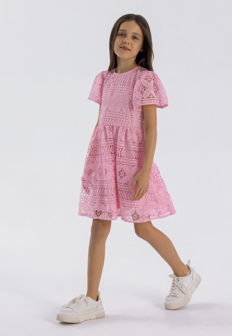 Rosa spetsklänning med korta ärmar, med intrikata mönster och en utsvängd kjol. Kombinerad med vita sneakers för en avslappnad look.