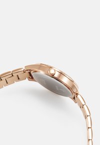 Armbanduhr aus roségoldfarbenem Metall mit rundem Gehäuse, glattem Lünettenrand, geriffelter Krone und einem Gliederarmband. Rückseite mit Markenlogo und technischen Details.