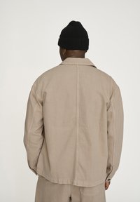 Beige Canvasjacke mit lockerer Passform, sichtbaren Nähten und Seitentaschen. Getragen mit einer schwarzen gerippten Strickmütze.