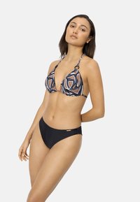 Mujer de pie con un top de bikini halter estampado y parte inferior de bikini negra lisa, contra un fondo claro y sencillo.