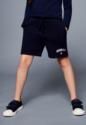 Enfant portant un short bleu marine avec le texte « DOUBLE Y » et un logo en forme de bouclier, associé à des baskets bleu marine avec semelles blanches et fermetures velcro.