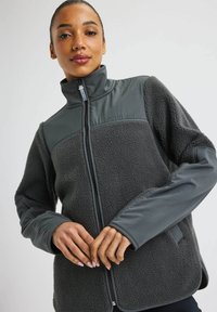 Tummanharmaa fleece-takki, jossa on erottuvat sileät kangasosat, korkea kaulus, täysipitkä vetskari ja rannekoristeet. Teksturoitu pinta logoyksityiskohdalla.