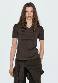 Bruine, kort-gevleugelde V-hals T-shirt gemaakt van een glad materiaal, gecombineerd met donkere gestructureerde broek en een geknoopte trui om de taille.