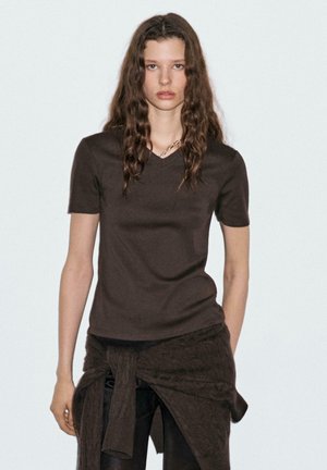 Brown, short-sleeve V-neck T-shirt made of a smooth material, paired with dark textured pants and a tied sweater around the waist.
