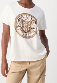 Bílý bavlněný tričko s grafikou motýla, zdobené složitými zlatými a růžovými detaily, obklopené dekorativními vzory. Volný střih.