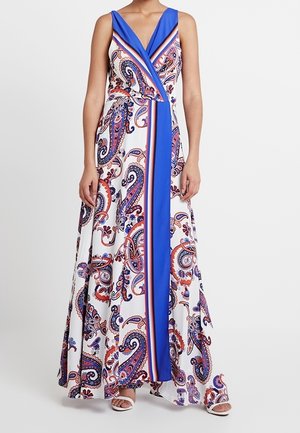 Ärmelloses Maxikleid mit blauem, weißem und rotem Paisley-Muster und einem einfarbigen blauen vertikalen Streifen, getragen mit weißen Sandalen mit offenem Zehenbereich.
