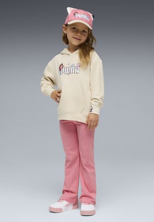 Dziewczyna w kremowej bluzie z kapturem z kreskówkowym logo Puma, różowych spodniach, dopasowanej różowej czapce Puma i białoróżowych trampkach, stojąca na szarym tle.
