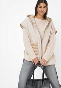 Beige hoodie vest met zachte tan accenten, gecombineerd met een lichte trui. Grijze jeans en een zwarte handtas maken de look compleet. Over het geheel een zachte textuur.