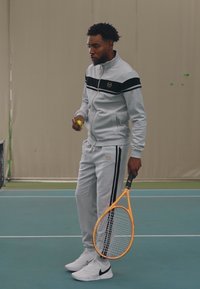 Grå atletisk treningsdrakt med svarte striper; holder en lys gul tennisball og en oransje tenniskølle; hvite sneakers. Innendørs tennisbane.