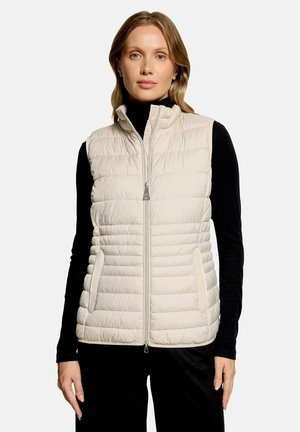 Femme portant un gilet matelassé sans manches beige clair sur un haut noir à manches longues, debout devant un fond clair uni.