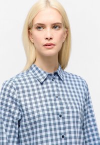 Jongeman met blond haar, draagt een blauwe en witte geruite blouse, kijkt iets opzij tegen een witte achtergrond.