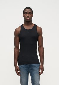Homem musculoso vestido com uma t-shirt justa preta e jeans azuis, em pé, de frente, contra um fundo claro e liso.