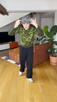 Pulover verde tricotat cu model abstract galben, pantaloni negri și șapcă camuflaj gri cu logo roșu; pantofi gri; decor interior cu plante.