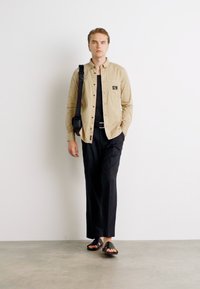 Camicia button-up beige con taschino, canottiera nera, pantaloni neri e sandali neri. Tessuto morbido e vestibilità casual.