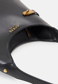 Sac en similicuir noir à texture lisse, doté d'une fermeture à pression dorée et du logo "GUESS" embossé. Le design comprend une bandoulière incurvée.