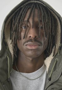 Olivgrüne Kapuzenjacke mit einem strukturierten Innenfutter, graues T-Shirt darunter und eine silberne Kette. Lange, dunkle Haare sind zu Dreadlocks frisiert.