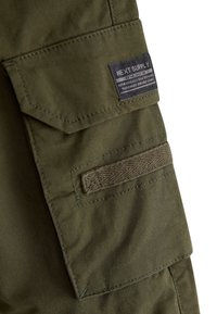 Gros plan sur une poche cargo en tissu vert olive avec rabat en Velcro et étiquette noire "Next Supply" cousue sur la partie supérieure.