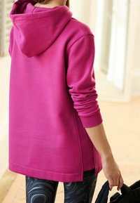 Fuchsiafarbener Hoodie mit weiter Passform, ausgestattet mit Kapuze, gerippten Bündchen und einem längeren Rückenteil. Kombiniert mit schwarzen, gemusterten Leggings.