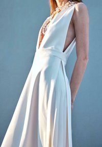 Femme portant une robe blanche sans manches avec un profond décolleté en V et un collier de perles sur un fond bleu uni.